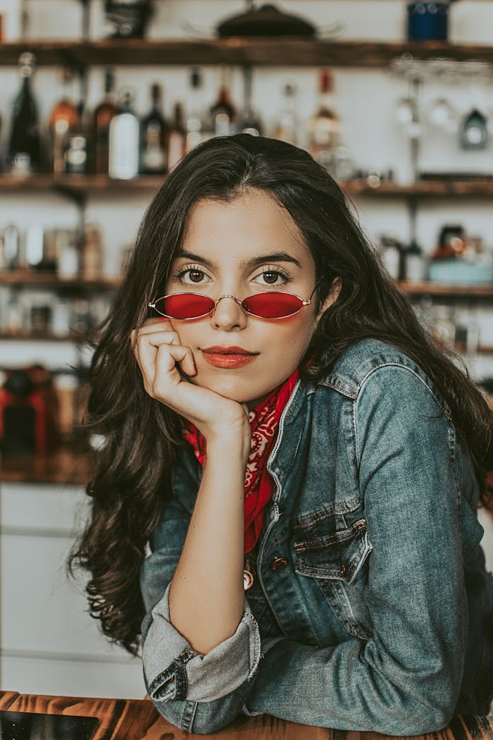 Fashionable woman with red sunglasses and denim jacket posing indoors at a bar.